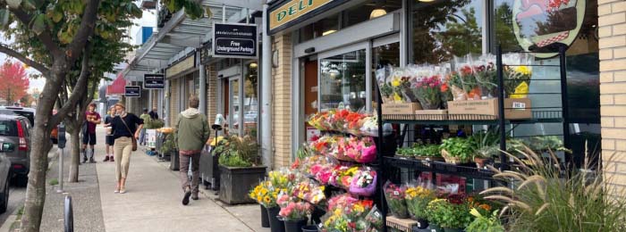 People walking along a tree-lined commercial street with an outdoor flower display.